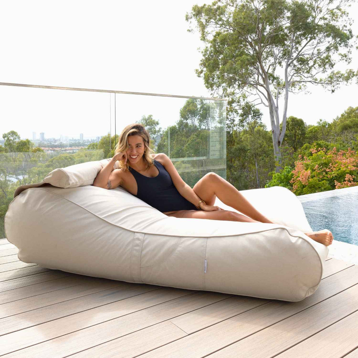Woman reclining on a Mooi Living Natural Nevaeh double outdoor bean bag sunbed on a timber deck beside a pool, MOSTNEVBB-BEIGE