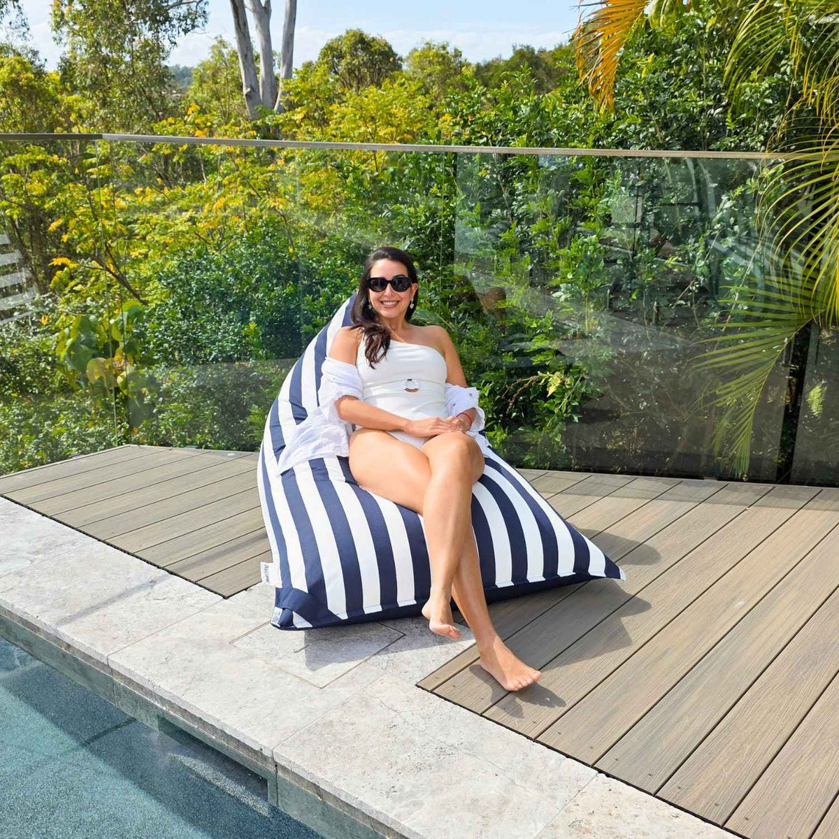 Wide view of a woman sitting on the navy and white striped side of a Mooi Living reversible triangle outdoor bean bag beside a pool, MOREVSTRIBB-NAVY