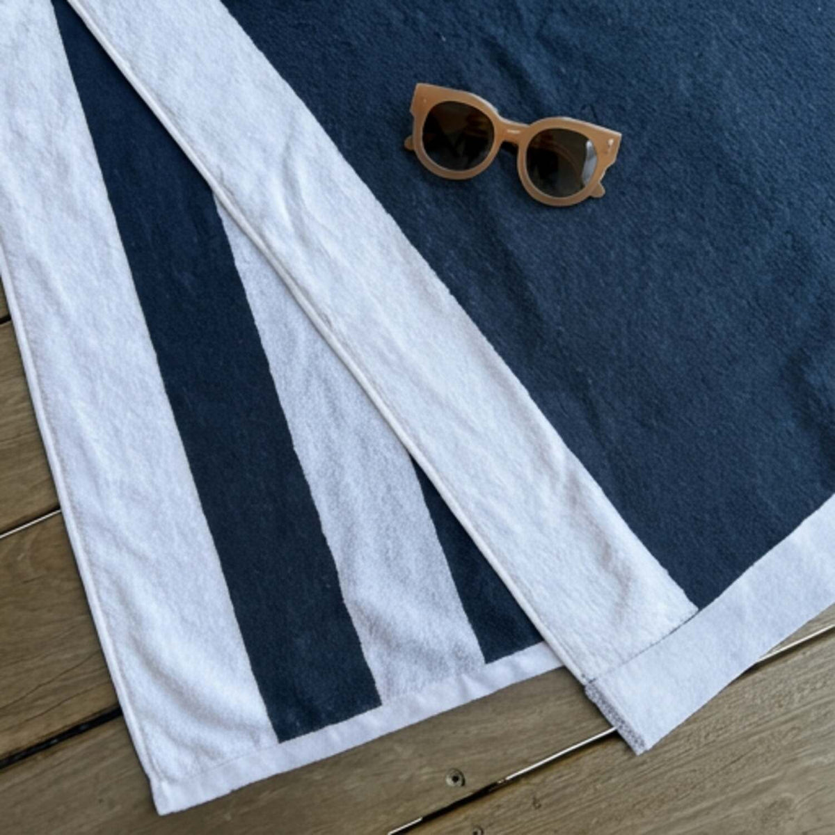 A navy blue and white striped beach towel by Mooi Living laid flat on a wooden surface, with a pair of round tan sunglasses resting on the towel.