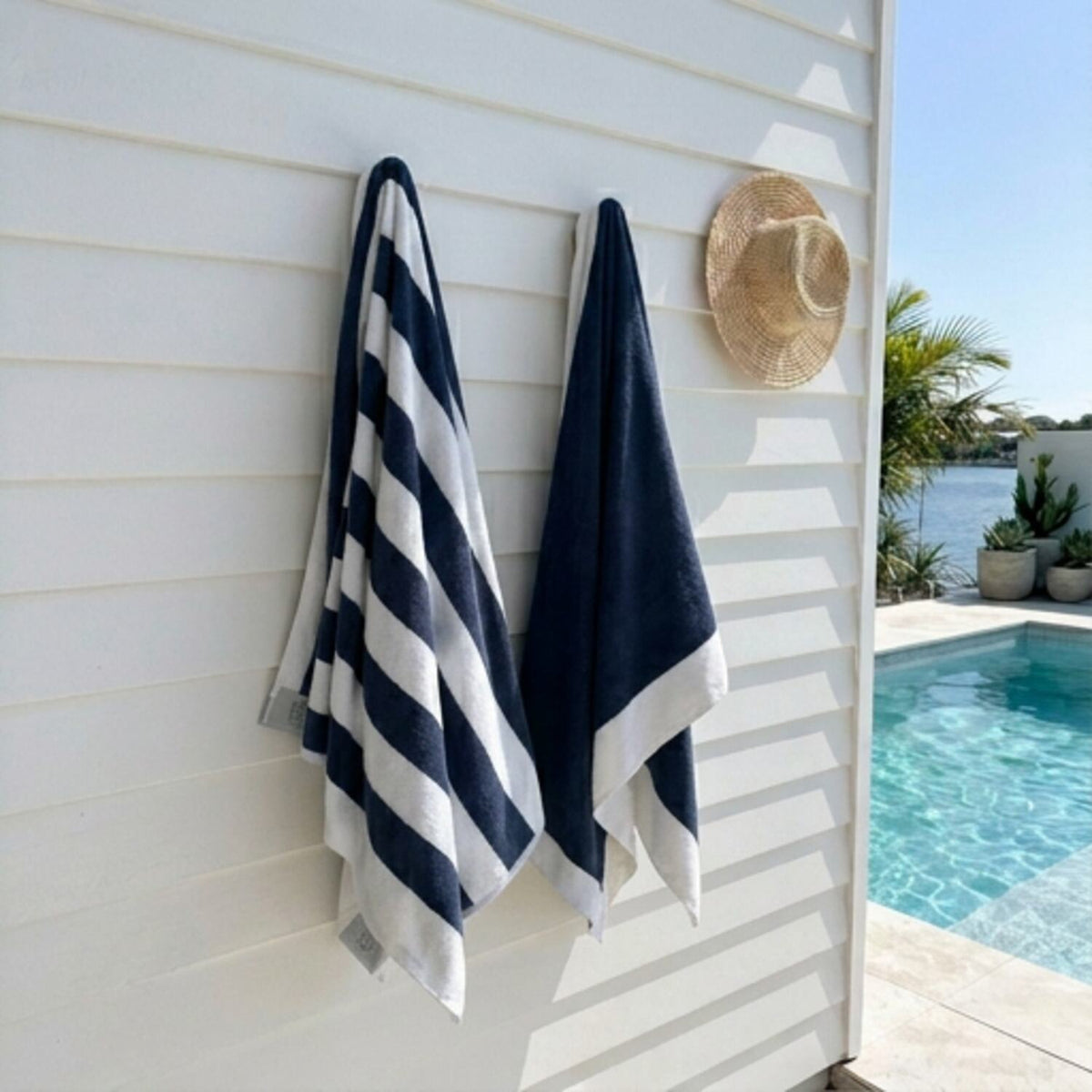 Two navy blue and white striped towels by Mooi Living hanging on a white exterior wall beside a pool, with a straw hat on the wall and a poolside background visible.