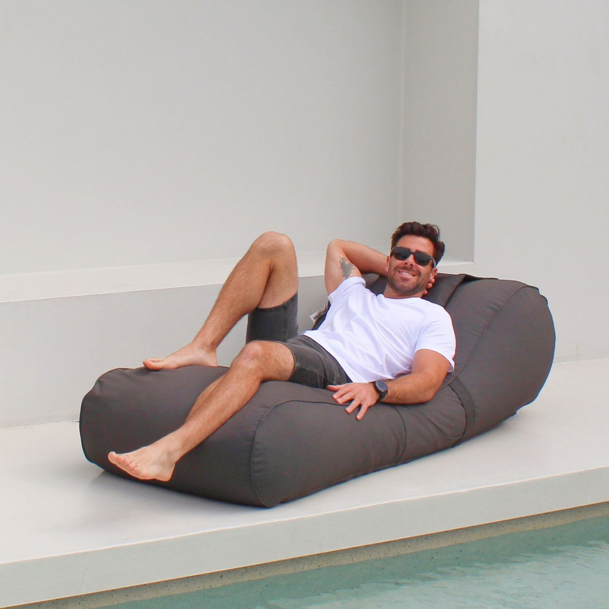 Man relaxing with one knee raised on a Mooi Living Oyster Black Isla outdoor bean bag sunlounger beside a pool, MOISLABB- OYSB