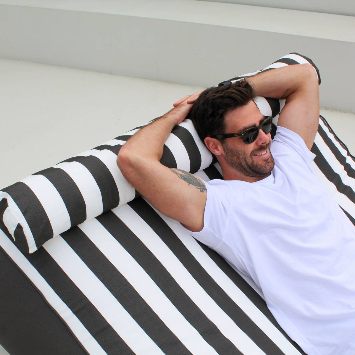 Close-up of a man reclining on a Mooi Living Oyster Black Nevaeh outdoor bean bag sunbed with a striped bolster cushion, MOSTNEVBB-OYSBLA
