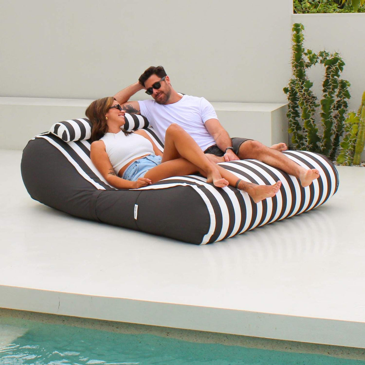 Couple relaxing on a Mooi Living Oyster Black Nevaeh double outdoor bean bag sunbed on a white patio beside a pool, MOSTNEVBB-OYSBLA