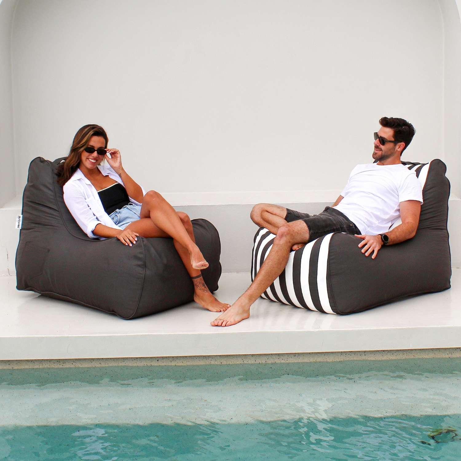 A man and a woman relax on Mooi Living Bora Bora bean bag chairs beside a swimming pool, MOBORABB-STRI-OYSBLA