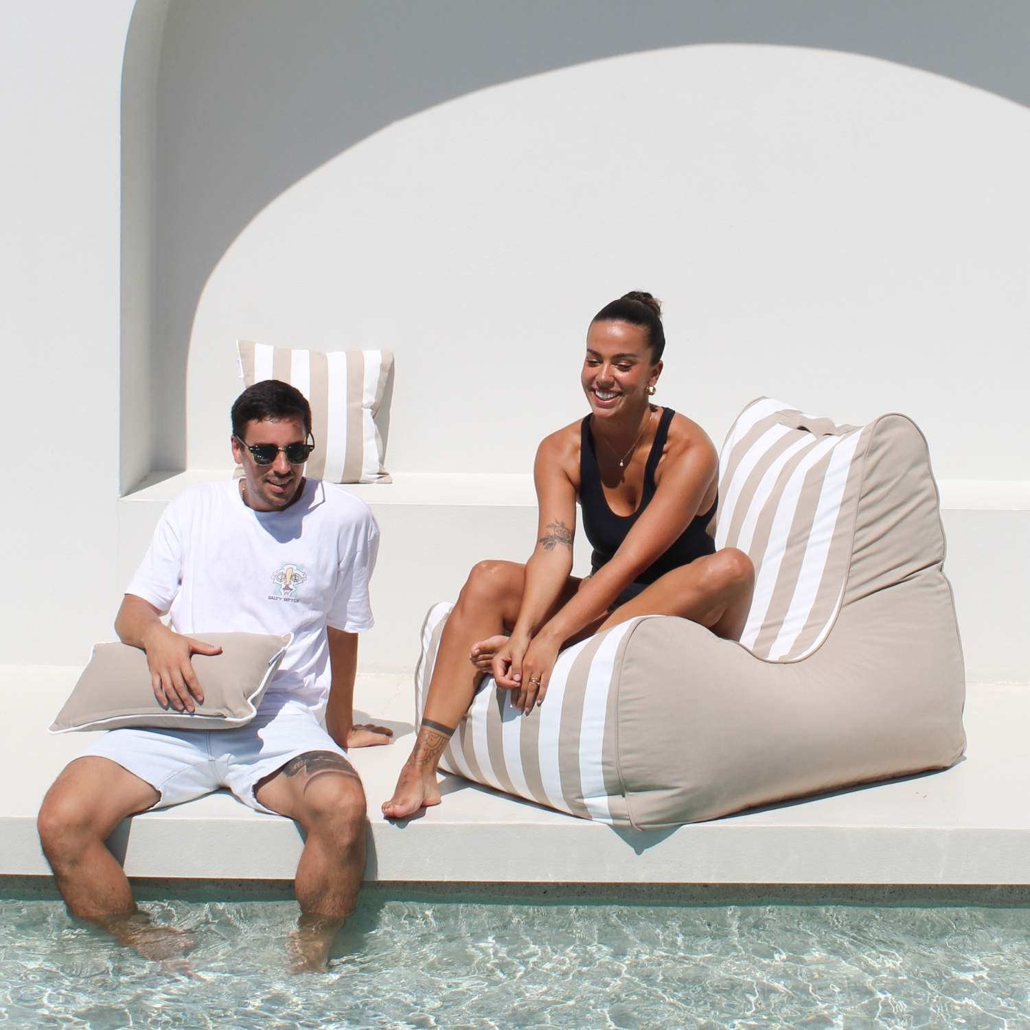 A man and a woman relax by a poolside on oversized beige and white striped bean bag loungers, with a white modern architectural background, MOBORABB-STRI-TAU