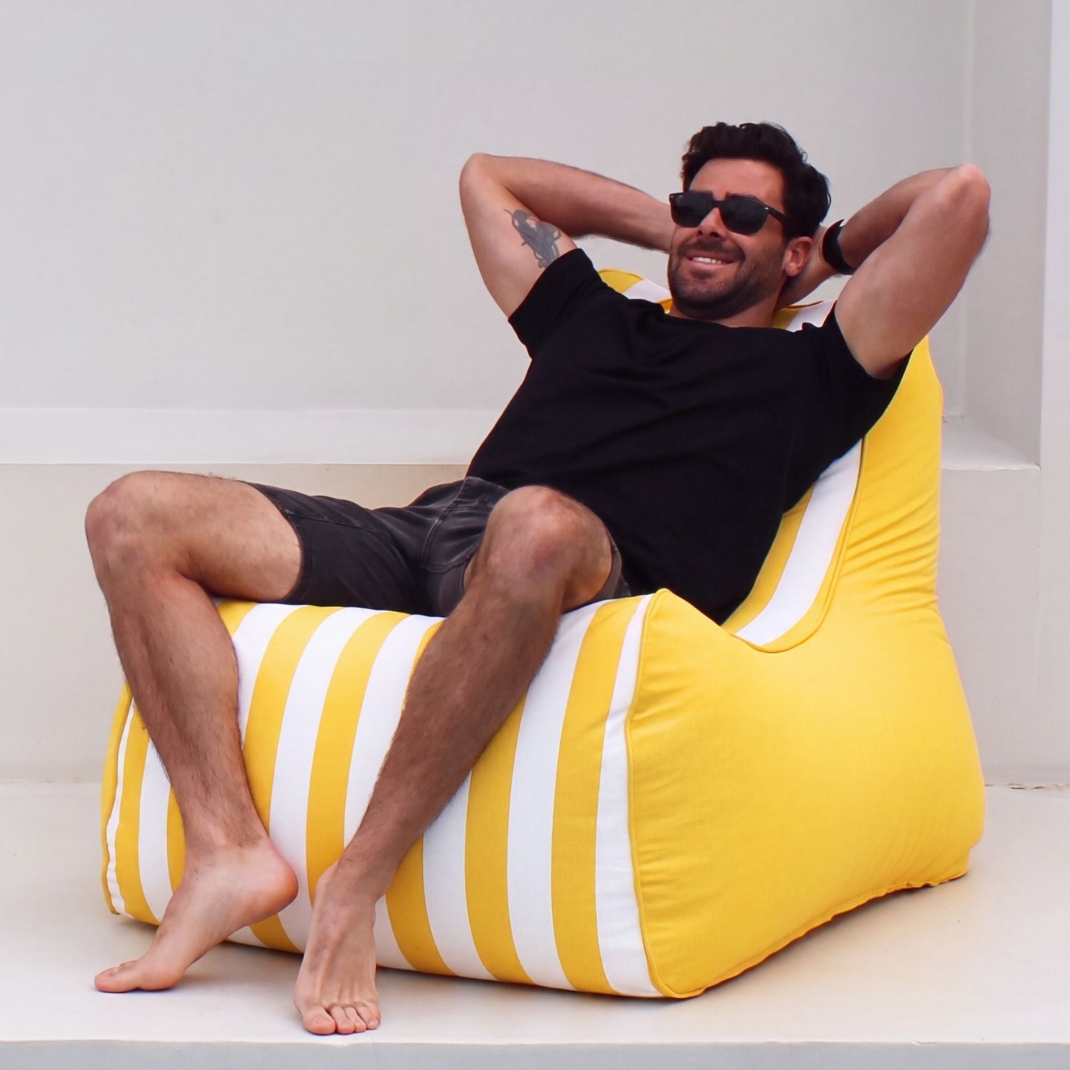 A man wearing sunglasses and a black t-shirt relaxes reclining on a yellow and white striped Bora bean bag chair against a clean white wall, MOBORABB-STRI-YEL