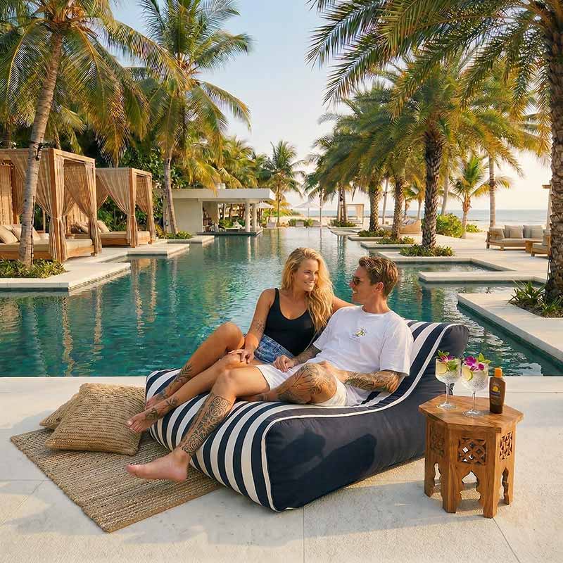 Luxury tropical resort pool with palm trees, white stone pool deck and turquoise water, two adults relaxing together on a Navy Stripe Chill UV double seater bean bag lounger beside the pool, drinks on a small side table, sunny holiday atmosphere.