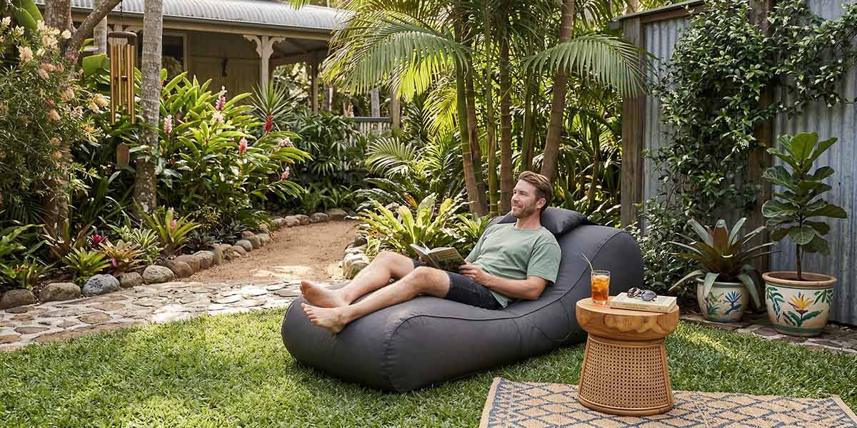 Australian Tropical backyard - Lush green garden with grass, trees and soft landscaping, Oyster Black Isla sunlounger Bean Bag placed on the lawn in dappled sunlight.