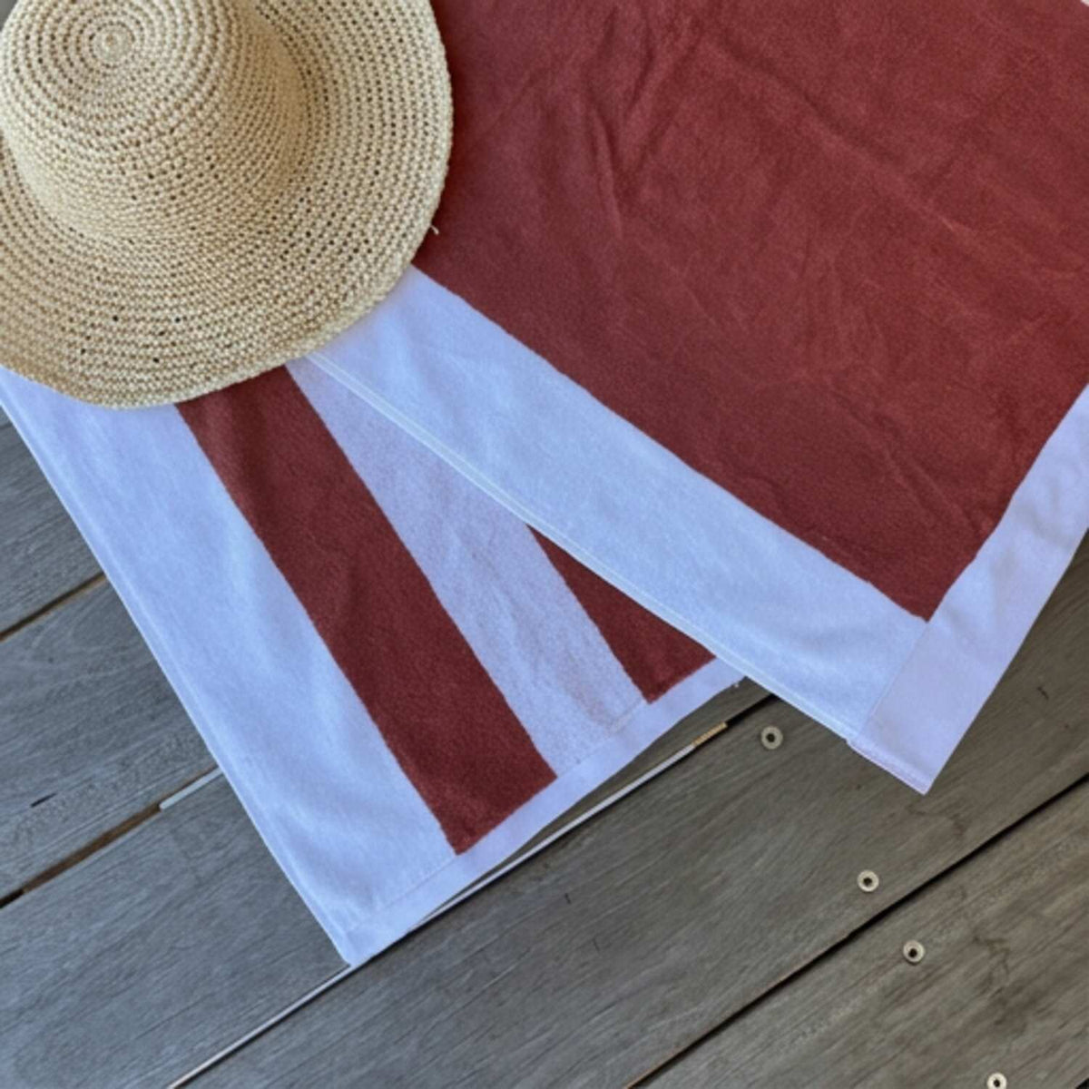 A terracotta red and white striped beach towel by Mooi Living laid out on weathered gray wooden planks, with a woven straw sun hat resting on top.