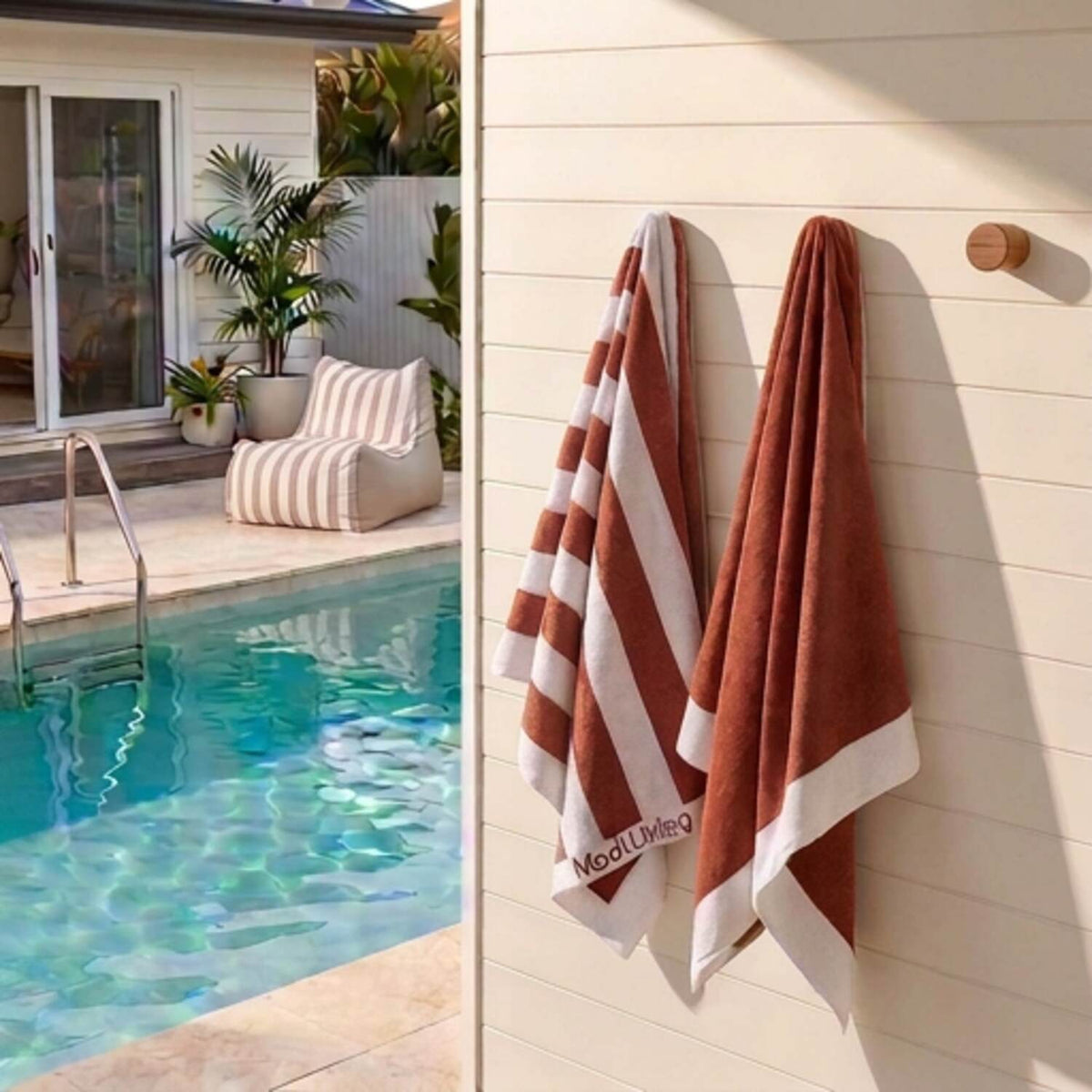 Two terracotta red and white striped towels by Mooi Living hanging on a cream-colored wall next to a swimming pool, with a straw hat on a hook above. A matching striped lounge chair and house exterior are visible in the background.