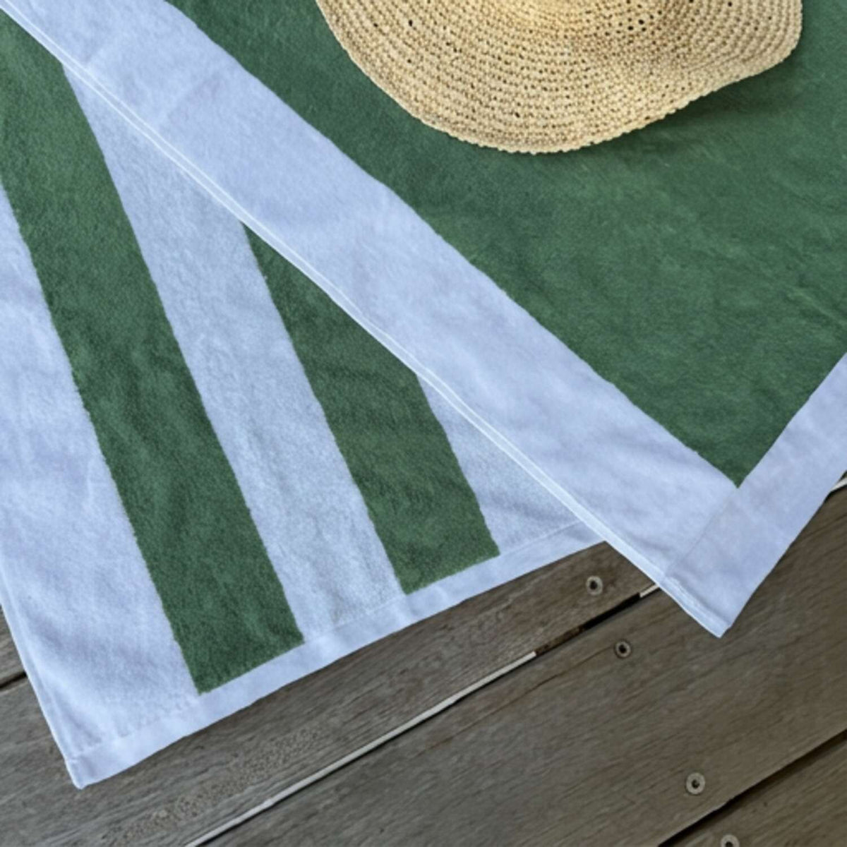 A green and white striped beach towel by Mooi Living laid on a wooden surface, with a woven straw hat partially visible at the top.