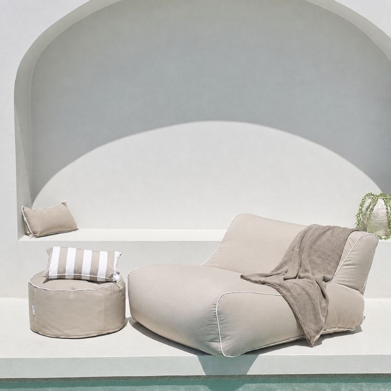 A Chill UV Lounger in Taupe with a matching Outdoor Ottoman, striped accent pillow, styled with a draped throw blanket in front of a white arched wall.