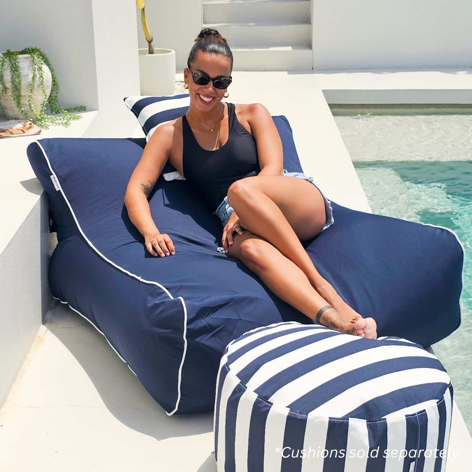 A woman wearing sunglasses reclines on a large navy Chill UV lounge bean bag by Mooi Living next to a swimming pool, with a matching striped pouf in front. The setting is a bright, modern outdoor patio, MOCHUVBB-NAVY.