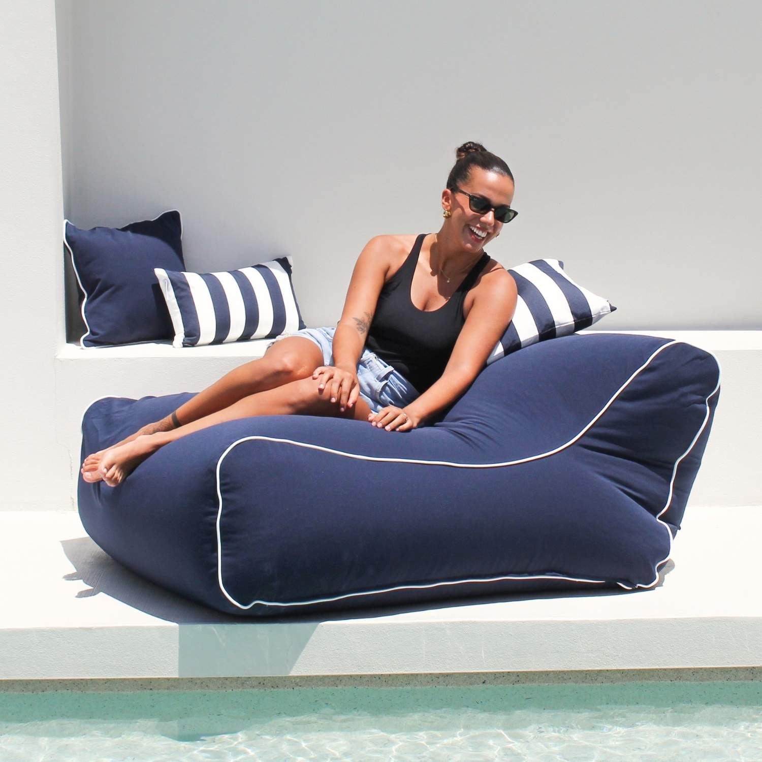 A woman wearing sunglasses sits on a large navy Chill UV outdoor bean bag chair with white piping by Mooi Living, MOCHUVBB-NAVY.