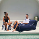 A man and a woman relax by a poolside. The man is reclining on a large navy blue Chill UV outdoor bean bag chair with white piping, while the woman sits on a striped navy and white pouf, MOCHUVBB-NAVY