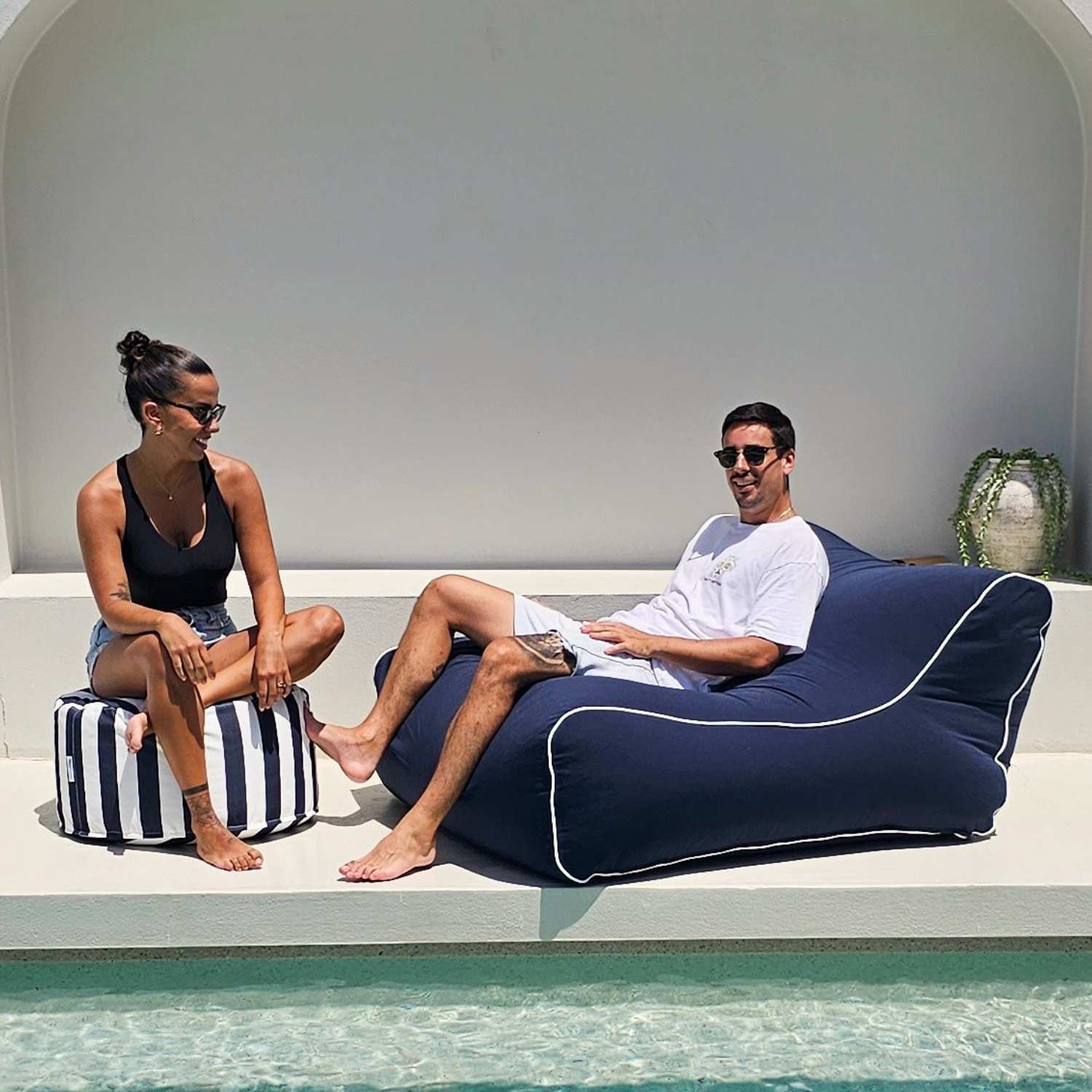 A man and a woman relax by a poolside. The man is reclining on a large navy blue Chill UV outdoor bean bag chair with white piping, while the woman sits on a striped navy and white pouf, MOCHUVBB-NAVY