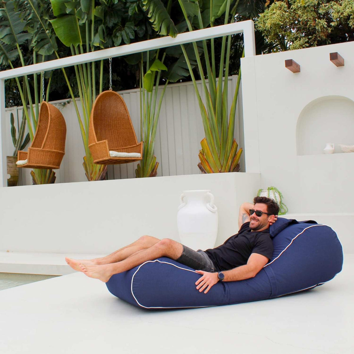 Wide side view of a man stretching out on a Mooi Living Navy Isla outdoor bean bag sunlounger on a patio surrounded by tropical plants, MOISLABB-NVY