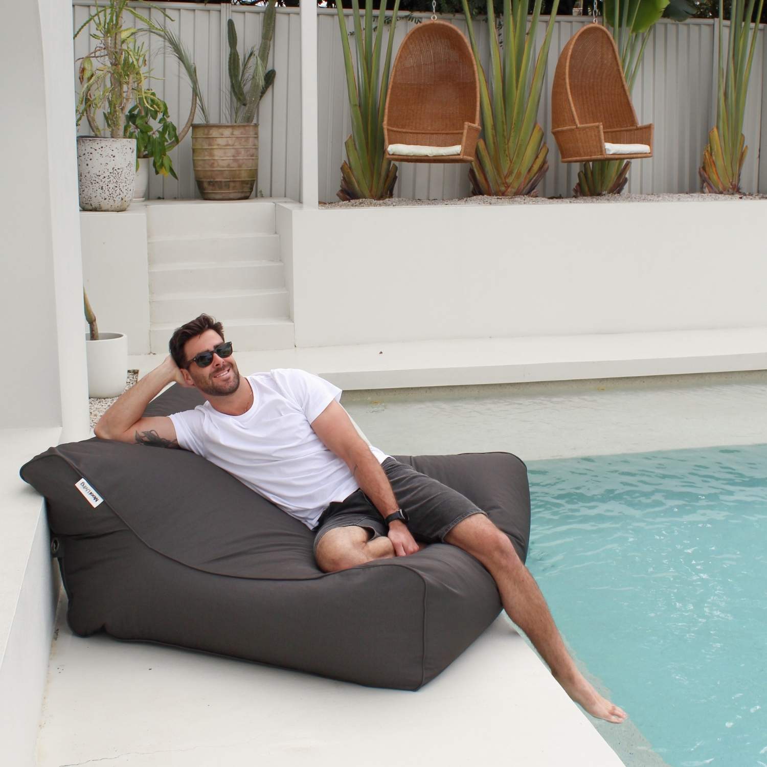 A man wearing sunglasses and a white t-shirt reclines on a Chill UV bean bag lounger next to a swimming pool, MOCHUVBB-OYSBLA