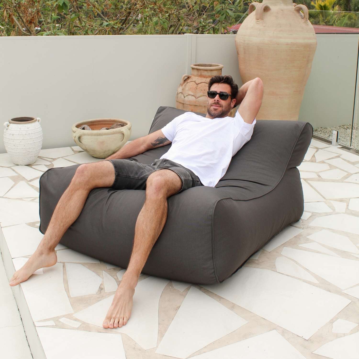 A man wearing sunglasses, a white t-shirt, and dark shorts relaxes reclining on a Mooi Living Oyster Black Chill UV bean bag chair, MOCHUVBB-OYSBLA.