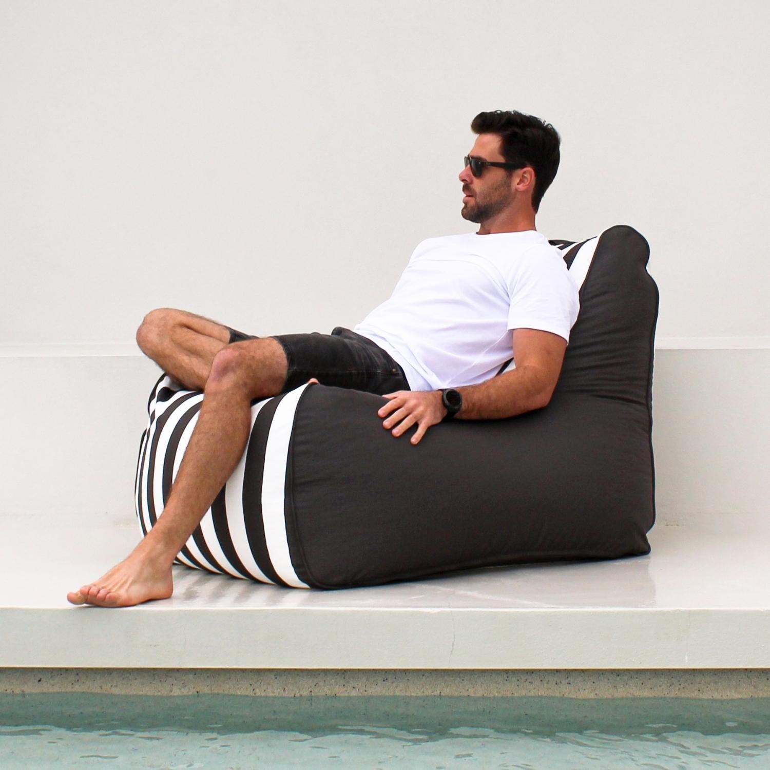 A man wearing sunglasses, a white t-shirt, and dark shorts reclines on a Mooi Living Oyster Black Stripe Bora Bora bean bag chair, positioned next to a swimming pool against a clean white wall,MOBORABB-STRI-OYSBLA
