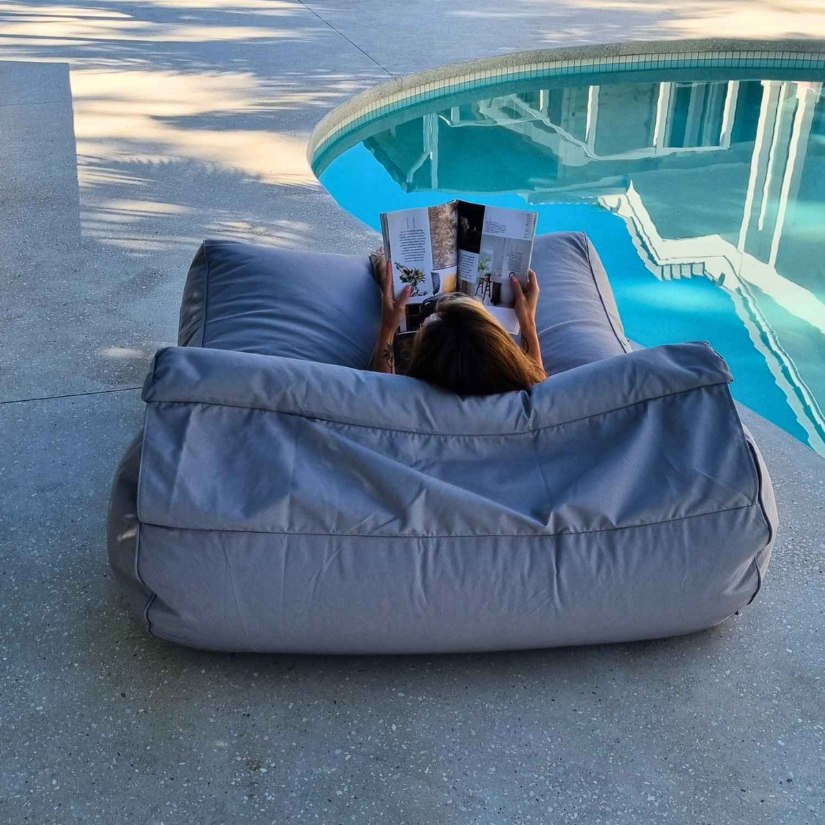Rear view of a woman reading on a Mooi Living Pumice Stone Nevaeh double outdoor bean bag sunbed, showing its backrest and bolster cushion beside a pool, MOSTNEVBB-PUMSTO