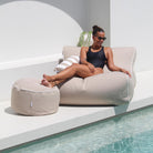 A woman in a black swimsuit and sunglasses relaxes on a taupe Chill UV outdoor lounger next to a pool, with a matching round ottoman and a striped pillow in a modern white patio setting, MOCHUVBB-TAUP