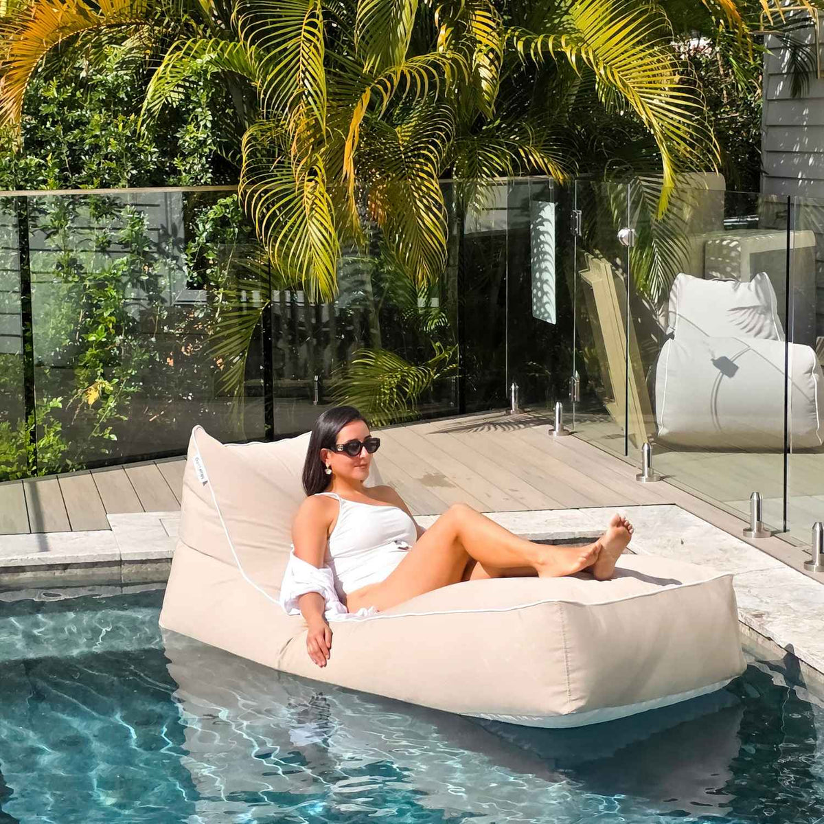 Woman relaxing on a Taupe Talia bean bag pool float beside a timber deck and tropical palms, MOTALFLTBB- TAUP