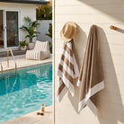 Two beige and white striped towels by Mooi Living hanging on a cream-colored wall next to a swimming pool, with a straw hat on a hook above. A matching striped lounge chair and house exterior are visible in the background.