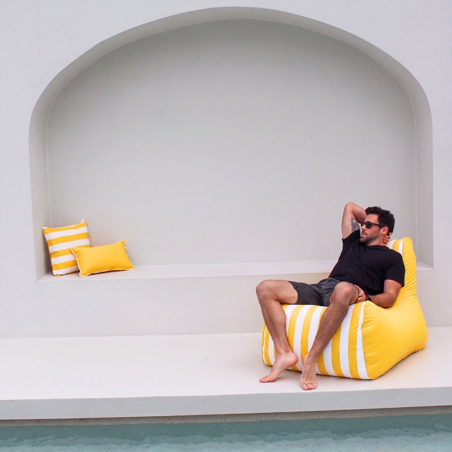 A man wearing sunglasses and a black t-shirt relaxes on a bright yellow and white striped Bora bean bag chair next to a pool, with a matching yellow and white striped pillow on a white bench in the background, MOBORABB-STRI-YEL