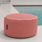 A Blush Outdoor Ottoman with white piping and a small black 'Mool Living' tag, positioned next to a swimming pool on a light concrete patio.