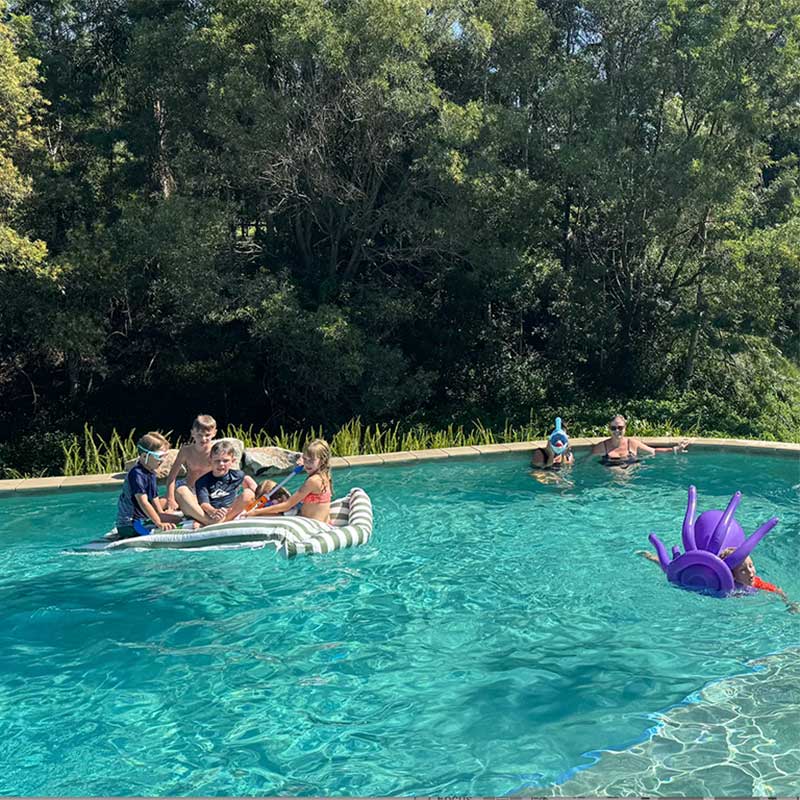 People enjoying a pool day with inflatable toys and children playing on a Santorini Floating Outdoor Bean Bag by Mooi Living, surrounded by trees.