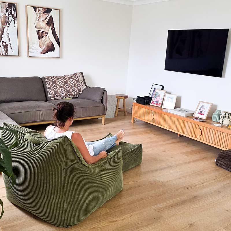 Cord Chair in Olive Green in a living room with a woman sitting on the corduroy bean bag chair. This bean bag is perfect for any indoor space.