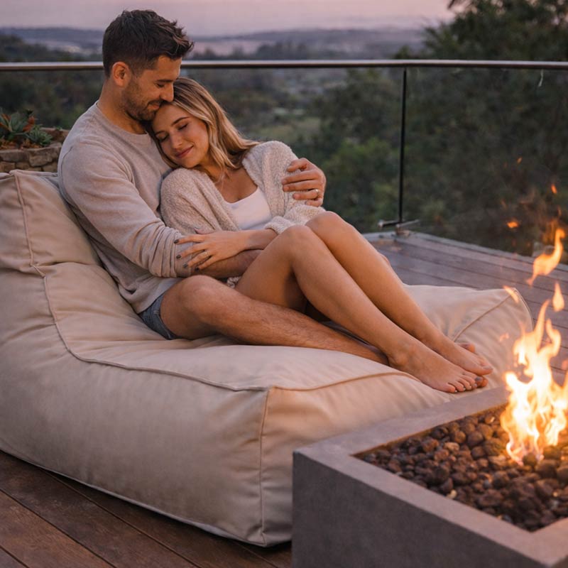 Lifestyle Image of the Natural Beige Lana Lounger with a couple sitting on the lounger bean bag by a fire pit with a scenic background