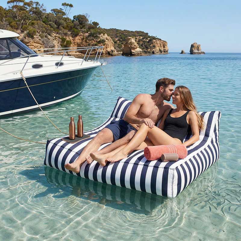 Lifestyle Image of the Navy Stripe Lana Lounger floating in the water, tied to a boat with two models lounging comfortably. 